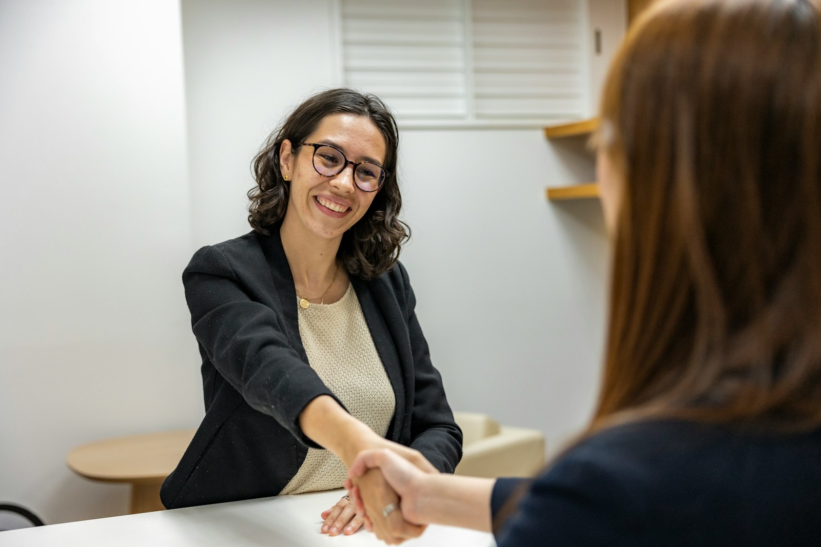 A woman in a black blazer smiles while shaking hands with another person, indicating a successful meeting or interview context.