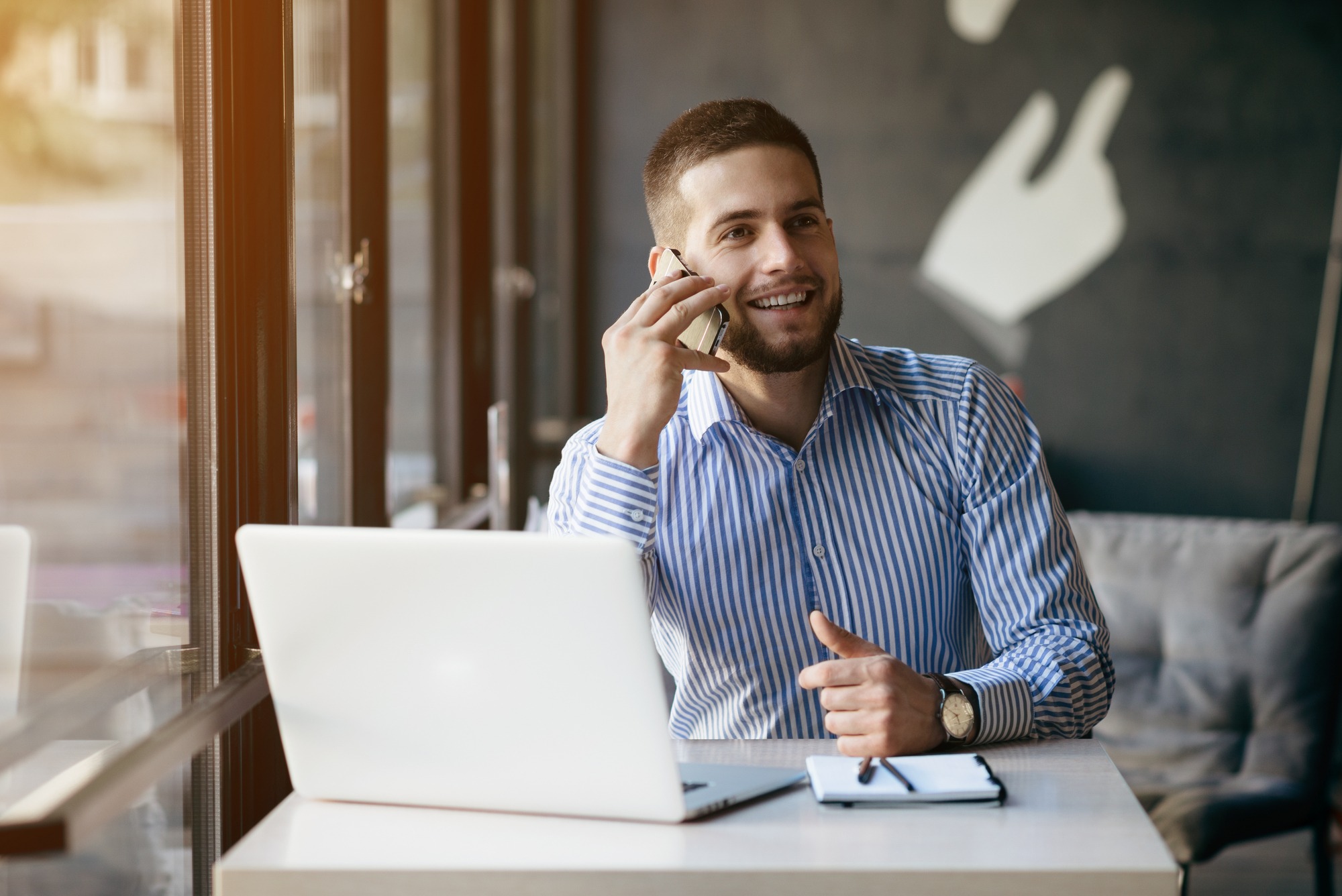 Smiling man in a striped shirt talks on the phone while sitting at a desk with a laptop and notepad, suggesting a productive work environment.